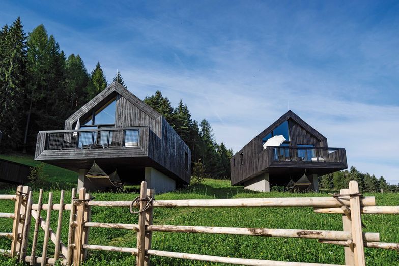 Die drei Chalets am Waldrand sind aus dunklem Holz und verschmelzen nahezu mit dem Wald dahinter. Ihre Geometrie und die einfachen gestalterischen Elemente nehmen Bezug auf die traditionelle Bauweise in der Region.